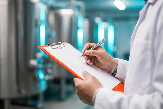 Close up of scientist hand writing on a blank clipboard for quality control in a brewery or pharmaceutical factory with stainless steel tanks background