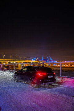 Moscow, Russia - December 13, 2024: Rear three-quarter view of a blue Infiniti QX50 crossover parked on snow with the illuminated yellow and blue ZSD bridge complex in the dark background