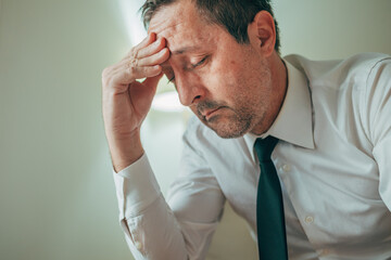 Fototapeta premium Troubled, exhausted businessman with a hand on his forehead, feeling overwhelmed by stress and fatigue.