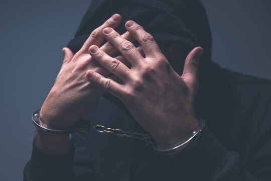 Low-key portrait of a hooded criminal with hands raised to an obscured face, handcuffs visible and dramatic shadows emphasizing tension and guilt.