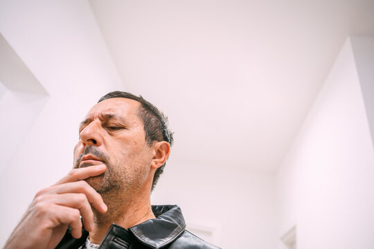 Mature man in leather jacket standing indoors, touching his chin with a thoughtful, contemplative expression. - Powered by Adobe