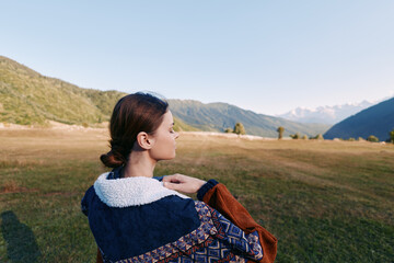 Fototapeta premium Woman in a warm jacket with fleece collar stands in a wide meadow overlooking distant mountains, profile view outdoors. Travel portrait capturing landscape, calm horizon and soft autumn light.