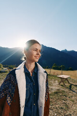 Fototapeta premium Woman portrait in mountains at sunrise with sunlight flare, smile and warm jacket over denim shirt. Outdoors field scene with calm rural landscape and soft backlight.
