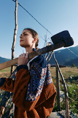 Fototapeta premium woman sledgehammer mallet rural vineyard mountains farmer outdoor carrying heavy wooden tool on shoulder, confident young agricultural worker in patterned jacket among trellis and fields under blue