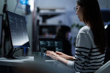 Rear view of a young programmer intensely reviewing code displayed on a monitor in a dark office. Software engineer typing and debugging on program. App development, hacker culture, and cybersecurity