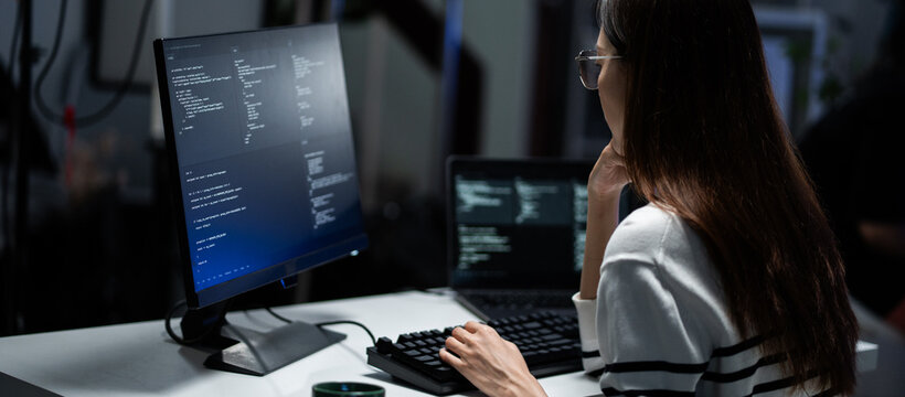 Rear view of a young programmer intensely reviewing code displayed on a monitor in a dark office. Software engineer typing and debugging on program. App development, hacker culture, and cybersecurity