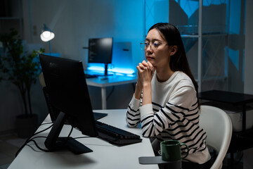 Serious Asian programmer wearing glasses pauses while reviewing code on the screen late at night. Software engineer in deep thought. Problem-solving, cybersecurity, remote work
