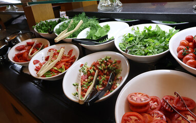 A colorful buffet of chopped tomatoes, cucumbers, peppers, herbs, and leafy greens arranged in white bowls with tongs, inviting guests to assemble a fresh, vibrant salad at a restaurant or event.