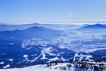 Panoramic winter view of Sheregesh ski resort in Russia see from snowy mountain Mystag slope. Snow-covered forest, village and hotels, distant peaks on horizon under clear blue sky and bright sun.