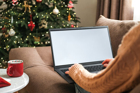 Woman in warm sweater is using laptop with blank white screen mock-up, sitting on sofa next decorated Christmas tree. Holiday online shopping, cozy winter digital lifestyle