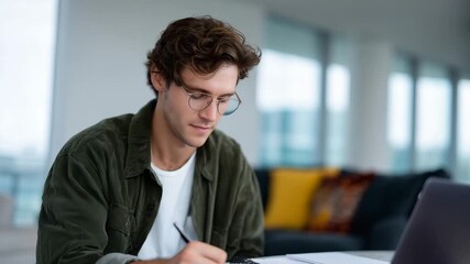 4k focused young businessman working from home at a wooden table, wearing modern glasses and an olive-green overshirt over a white T-shirt. He is leaning slightly forward toward his