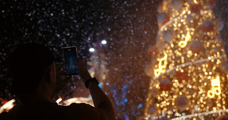 Silhouette of a person using a smartphone to capture the beautiful festive Christmas lights and bokeh at night. Represents holiday memories, celebration, and modern technology.