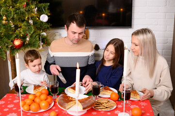 Mom, dad, and boy and girl are sitting at festive table, sharing Christmas meal and enjoying food. Family celebrates Christmas in warm, family atmosphere, by candlelight.