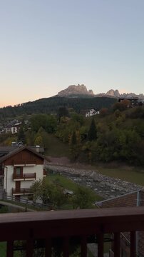 A scenic view of Moena, Italy, with mountains in the background, houses, and a street leading uphill during sunrise, showcasing an autumn landscape.