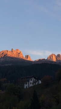 A breathtaking sunrise view in Moena, with the mountain peaks glowing in the early light, and a picturesque house nestled on the hillside, surrounded by lush green trees.