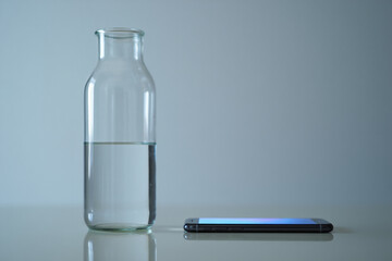 Glass Bottle Half Full of Water Near Modern Mobile Phone Placed on Table Inside Bright Room for Hydration Reminder and Digital Connection