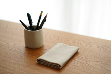Stationery Holder with Black Pens and Pencils Next to Folded Beige Napkin on Wooden Table Surface Near Window with Neutral Color Scheme