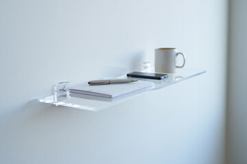 Clear Shelf with Phone, Mug, Notepad and Pen against White Wall in Minimalist Home Office Setting 150 characters