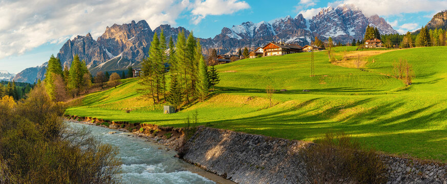 Panoramic landscape in Cortina d'Ampezzo with alpine river, green meadows, wooden houses and snow covered Dolomiti Alps mountains. Italian Dolomites in Belluno region, Italy
