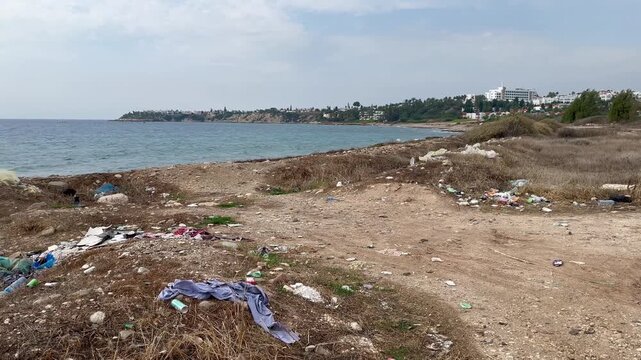 Trash-strewn coastal ground with scattered plastic waste and discarded items along a rocky shoreline, contrasting with clean distant buildings and highlighting environmental pollution in Cyprus