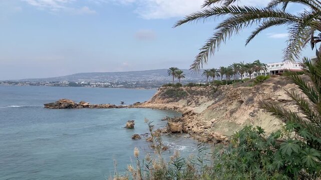 Rocky Mediterranean coastline with turquoise water, rugged cliffs and palm trees under a soft cloudy sky, capturing the calm coastal atmosphere and natural beauty of the Paphos surroundings in Cyprus