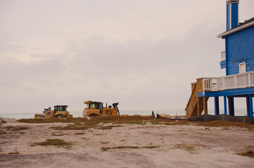 A partially elevated Beach House Damaged by Storm With Exposed Framework and Sand Underneath on the Shoreline in Treasure Island, FL. Sagging deck, and sand-filled ground. The scene captures coastal e