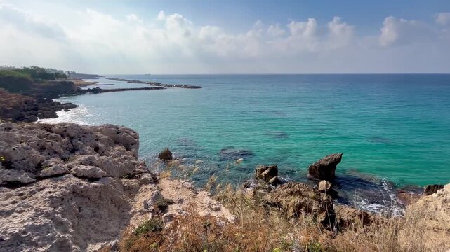 Rocky Mediterranean shoreline with turquoise waves and rugged stone formations under soft clouds, captured in a calm coastal scene with a clear view toward Alyki Beach and tranquil seaside atmosphere