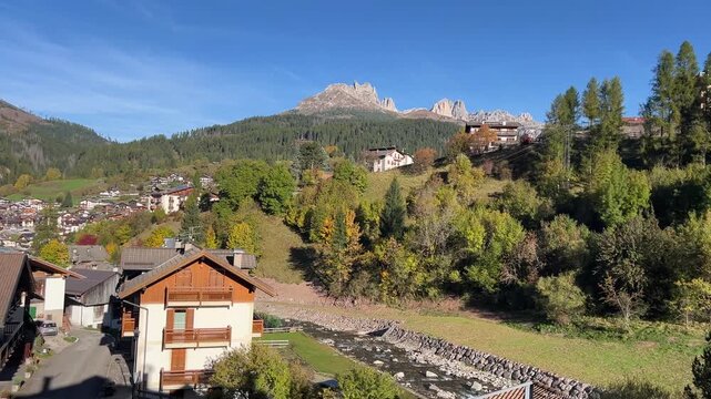 A beautiful village scene in Moena, located in the Dolomites, with traditional houses, green hills, and majestic mountain peaks under a clear blue sky.