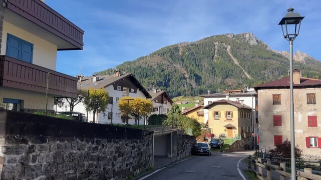 A charming street view in Moena, with traditional Italian houses lining the road, leading up to a towering mountain in the background, under a clear blue sky.