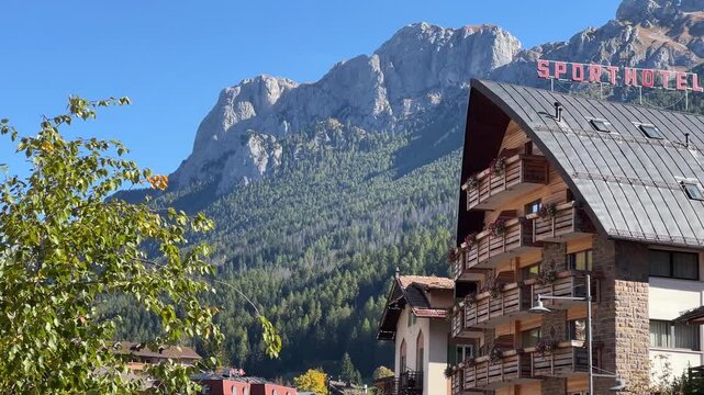 A close-up of a charming building in Moena, featuring the &ldquo;Sporthotel&rdquo; sign, with the stunning Dolomites in the background, surrounded by greenery and clear blue skies.