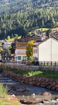A picturesque view of the town of Moena nestled in the mountains, with houses lining a river, surrounded by lush greenery and towering trees. 