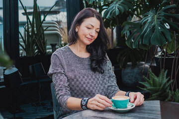 Middle-aged woman enjoying a cup of coffee in a cosy indoor garden, surrounded by lush greenery and natural light, creating a serene atmosphere with copy space