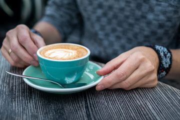 Woman holding a turquoise coffee cup with latte art, resting on a green saucer, on a rustic wooden table, enjoying a moment of relaxation and warmth in a cozy cafe atmosphere