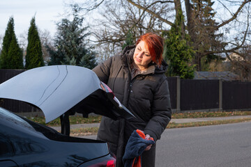 Woman with red hair wearing a black coat is opening the trunk of a black car on a sunny day, surrounded by trees and a suburban street, showcasing everyday life moments