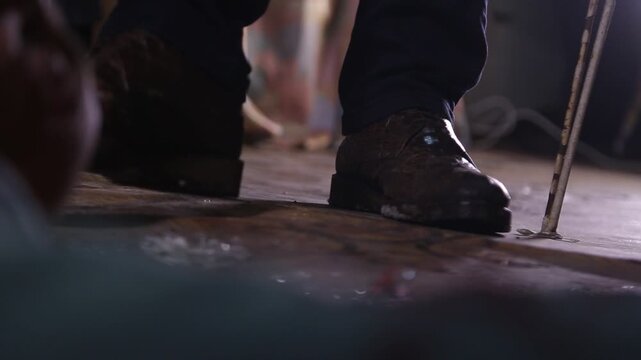 Close-up of feet in leather shoes stepping on broken glass shards on a dirty floor. Dark cinematic shot representing danger, abandonment, crime scenes, or the aftermath of destruction.

