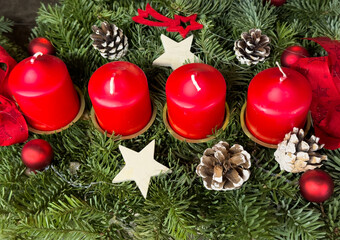 Advent wreath with four red candles, pine branches, cones and small Christmas decorations arranged in a traditional holiday design.