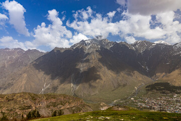 Vast mountain range with snow-capped peaks under a bright blue sky with fluffy white clouds. A small village nestled in a valley below, surrounded by rugged terrain and green slopes.