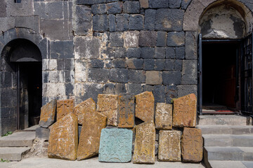 Sunlit ancient stone church with arched doorways, featuring a pile of weathered, ornate Armenian khachkars&mdash;carved crosses&mdash;against its dark, mossy stone wall.