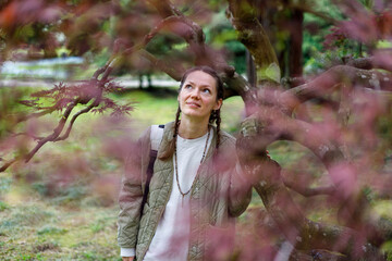 A woman with braided hair, wearing a quilted jacket and beaded necklace, gazes upward through blurred pink maple branches in a sunlit garden.