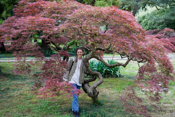 A smiling woman stands beside a vibrant red-leafed maple tree in a sunlit garden. Its twisted trunk and cascading foliage contrast with green grass and background trees under soft daylight.