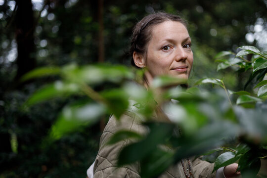 A woman with brown eyes and braided hair smiles softly, framed by vibrant green leaves in a lush, dimly lit forest. She wears a beige quilted jacket, with soft natural light filtering through the cano