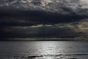 Dramatic, dark clouds loom over a vast, shimmering ocean. Faint sunbeams pierce the gloom, casting a soft glow on the water&rsquo;s surface under moody, low-light conditions.