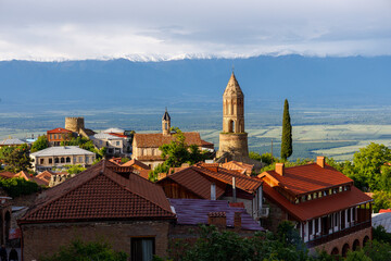 Sunlit village with terracotta roofs and stone towers, nestled against snow-capped mountains under a soft, cloudy sky. Lush greenery and expansive valleys stretch below.