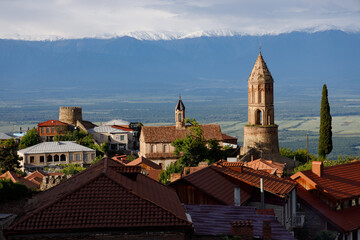 Sunlit village with terracotta roofs and stone towers, nestled against snow-capped mountains under a partly cloudy sky.