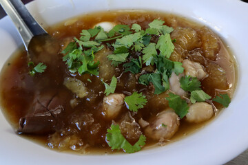 A close-up of a bowl of thick, brown, savory Fish Maw Soup containing fish maw, chicken meat,  dark coagulated blood, and topped with fresh coriander.