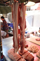 Two long strips of raw pork belly are hanging from hooks in a butcher or market stall, with other cuts of meat on a stainless steel counter with a blurry background.