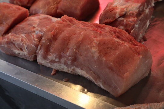 A large, fresh cut of raw pork loin is displayed on a shiny metal counter, likely at a butcher shop or market, with other cuts visible in the background.