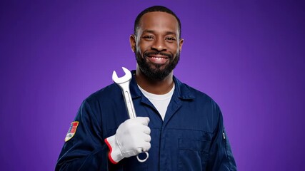 African American auto mechanic in a blue uniform and gloves posing with a wrench, looking cheerful. Handsome repairman in a workshop setting with a purple backdrop