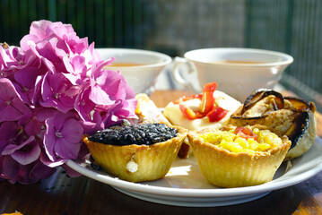 Tartlets filled with risotto and vegetables with pink hydrangea flowers beside tea cups on a rustic wooden table. Symbolizes afternoon tea elegance, garden party catering, and European bakery style.