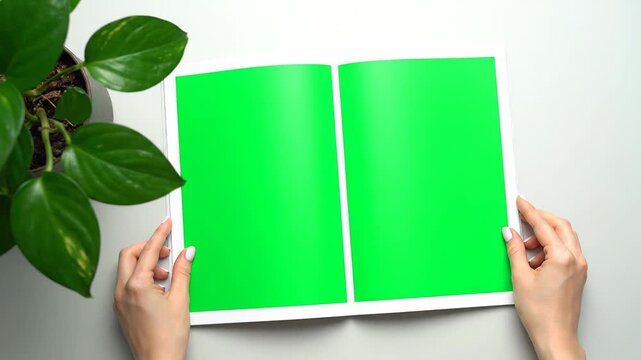 Woman opening a blank magazine with a green screen for content replacement. Top view of a person reading a book on a white office desk
